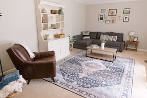 Un salon avec un fauteuil en cuir marron, un canapé gris, une table basse, un tapis à motifs et un meuble blanc avec des étagères.
