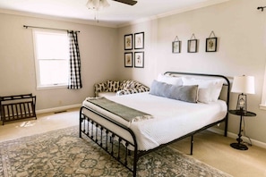Un dormitorio con cama, mesita de noche con lámpara, ventana con cortinas y un cuadro en la pared.