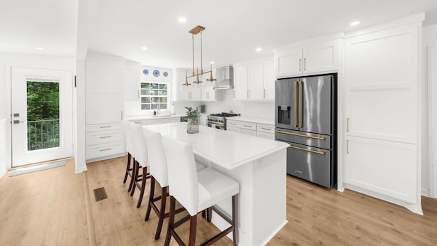 Una cocina moderna con armarios blancos, una isla central con taburetes altos y electrodomésticos de acero inoxidable, con vistas a zonas verdes exteriores.
