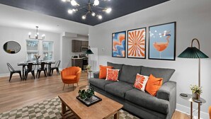 Een moderne woonkamer met een grijze bank, oranje kussens, een houten salontafel, en een eethoek met een zwarte tafel en stoelen.