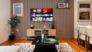 A modern living room with a flat-screen TV, a wooden cabinet, a coffee table with a Scrabble box, a patterned rug, and a dining area with chairs. - Boutique apartment near Old Trafford · terrace · sleeps 6 (Greater Manchester)