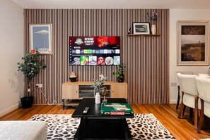 A modern living room with a flat-screen TV, a wooden cabinet, a coffee table with a Scrabble box, a patterned rug, and a dining area with chairs. - Boutique apartment near Old Trafford · terrace · sleeps 6 (Greater Manchester)