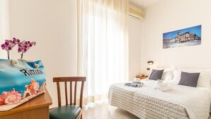 A hotel room with a bed, a chair, a desk with a bag, a window with curtains, and a wall-mounted picture. - Hotel Lariana (Rimini)