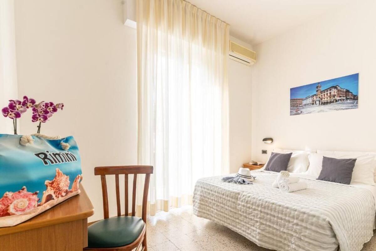 Ein Hotelzimmer mit Bett, Sessel, Schreibtisch mit Tasche, Fenster mit Vorhängen und einem Wandbild.