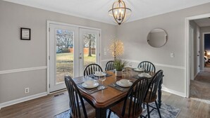 Un'area pranzo con un tavolo in legno, sedie nere, un lampadario e vista su una zona esterna attraverso porte in vetro.