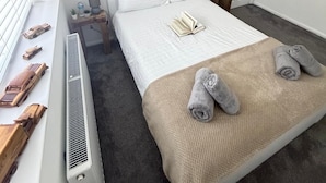 Et soveværelse med seng, radiator, et lille bord med lampe, en dør og et kunstværk på væggen.