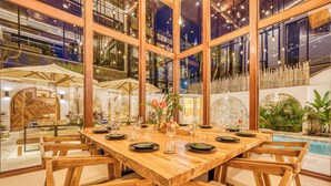 Un comedor con mesa y sillas de madera, rodeado de ventanas amplias y plantas de interior.