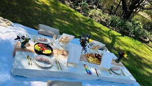An outdoor picnic setup with a table covered in a white cloth, featuring plates, cutlery, a fruit bowl, and a small vase with flowers. - Room-starling Room - Staynest Guesthouse (Johannesburg)