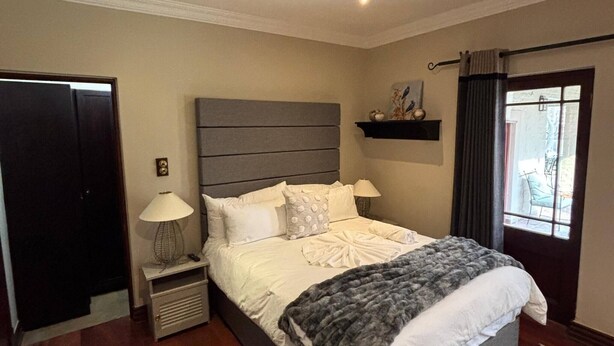 A bedroom with a large bed, bedside tables, a nightstand, a window with curtains, and a door. - Room-starling Room - Staynest Guesthouse (Johannesburg)