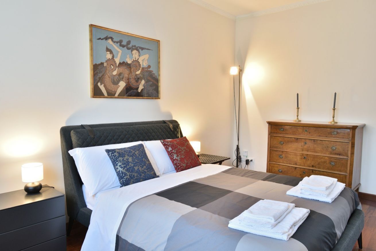 A bedroom with a bed, a nightstand, a dresser, a painting, and a floor lamp.