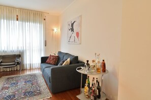 Un salon avec un canapé gris, un tapis à motifs, une petite table ronde sur laquelle sont posées des bouteilles, et une œuvre d’art murale.