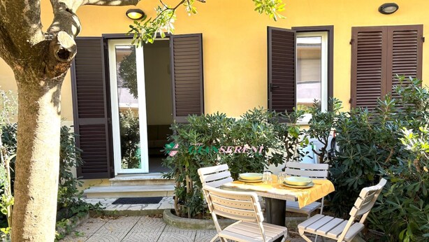 Un patio con una mesa, sillas, un pequeño árbol y un edificio amarillo con contraventanas.