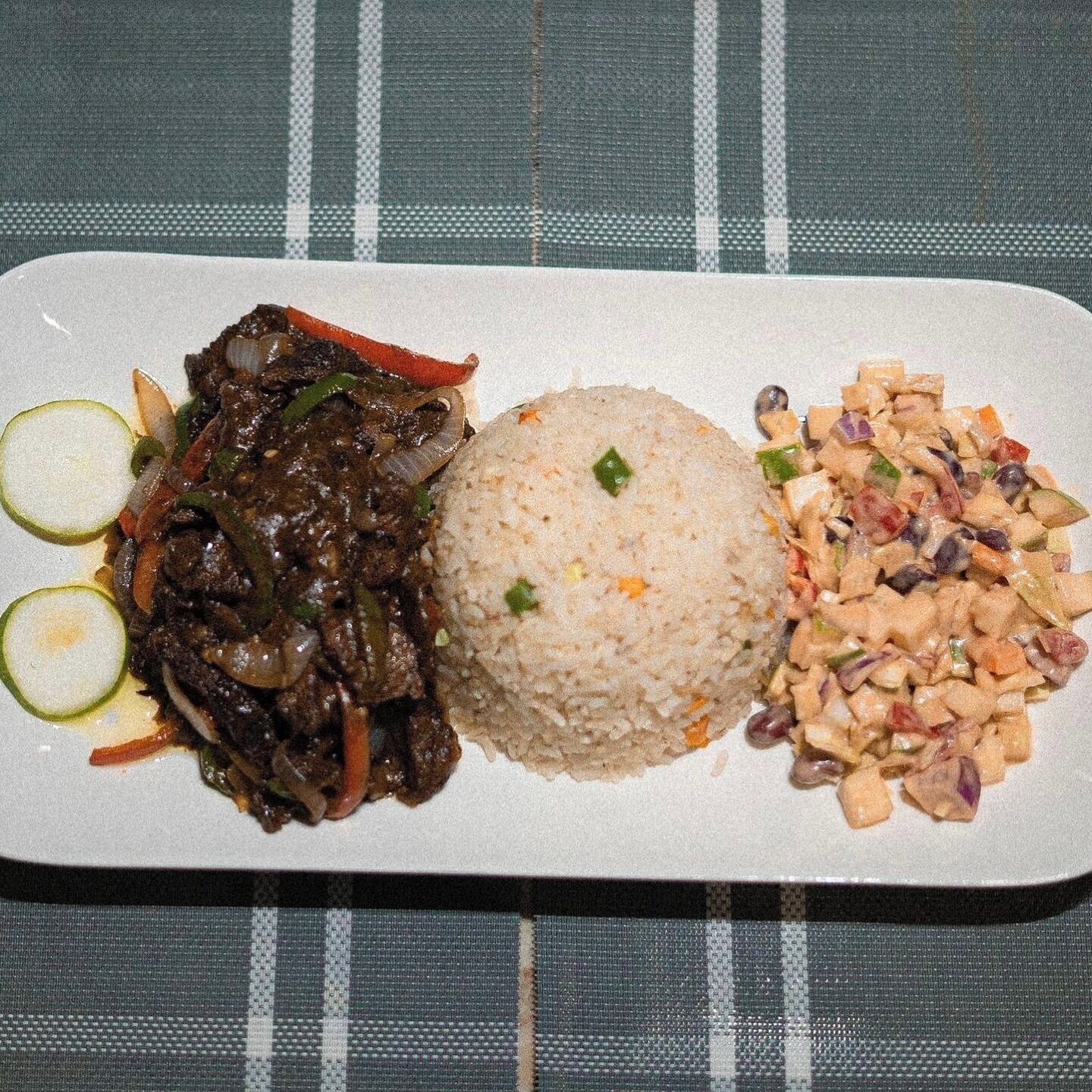 A rectangular plate with a meal consisting of stir-fried meat, rice, and a side of beans.