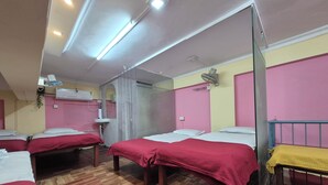 Habitación de hotel con cuatro camas, piso de madera, un espejo, un equipo de aire acondicionado y una mesita de noche con tela amarilla.