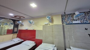 Habitación de hotel con tres camas individuales, una pared de acento roja, varios ventiladores de pared, un espejo grande y un salpicadero de azulejos.