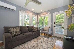 A living room with a grey sofa, a patterned throw pillow, a lamp, and a view through glass doors.