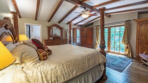 Ruime slaapkamer met een groot bed, houten balken, een plafondventilator en uitzicht op groen.