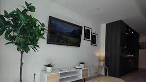 A modern living room with a TV, a plant, a small table, and a dark kitchen area. - Luxury Executive 1+Den | Office | Pool, Gym, Sauna | Free Parking (Toronto)