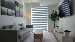 A modern living room with a grey sofa, a round coffee table, a TV mounted on the wall, and a large window with blinds. - Luxury Executive 1+Den | Office | Pool, Gym, Sauna | Free Parking (Toronto)