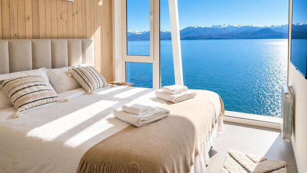 Una camera da letto con un'ampia finestra vista su un lago e montagne, un letto con cuscini e coperta, e rivestimenti in legno.