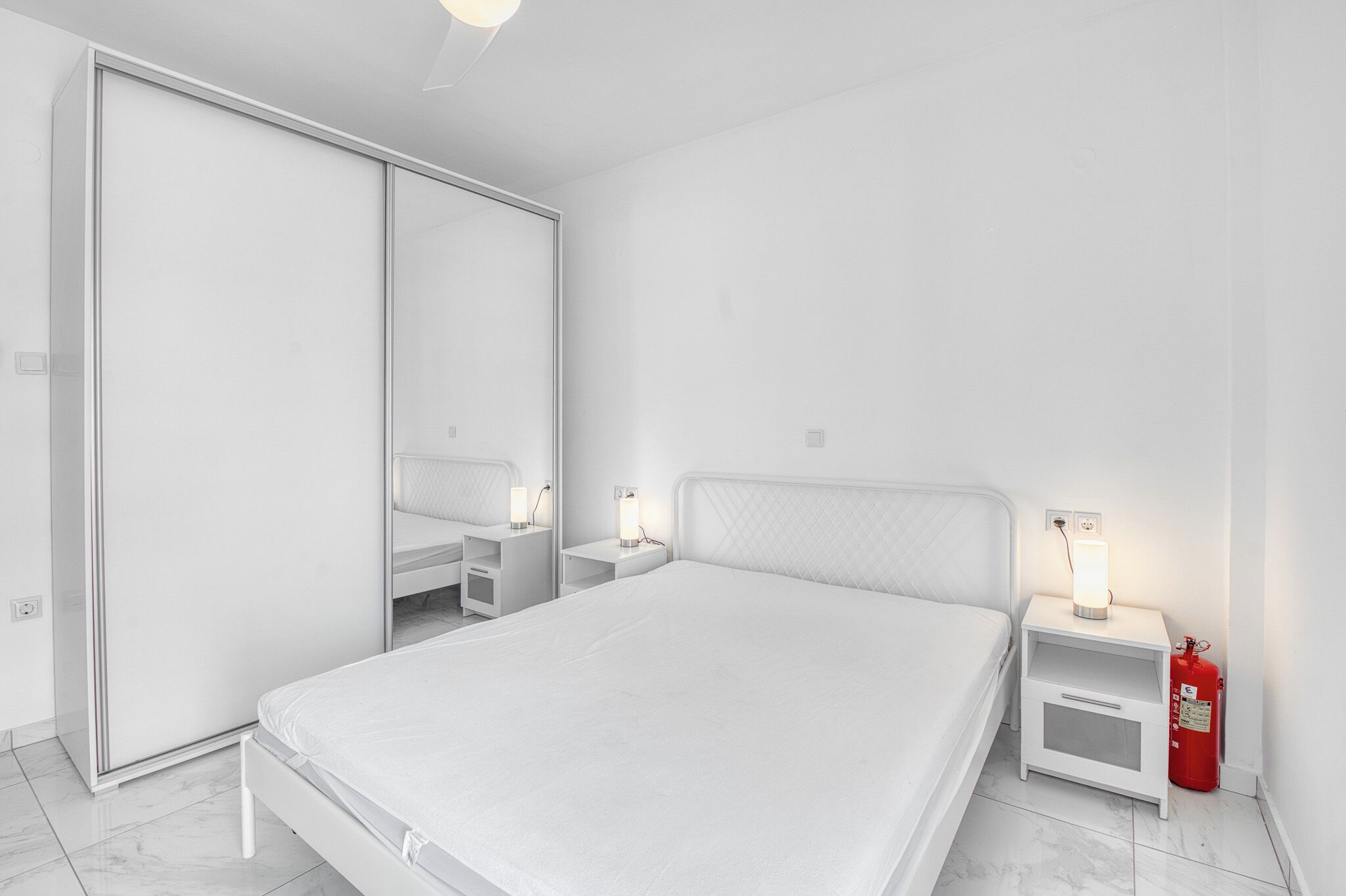 A bedroom with a bed, a wardrobe, a bedside table with a lamp, and a fire extinguisher.
