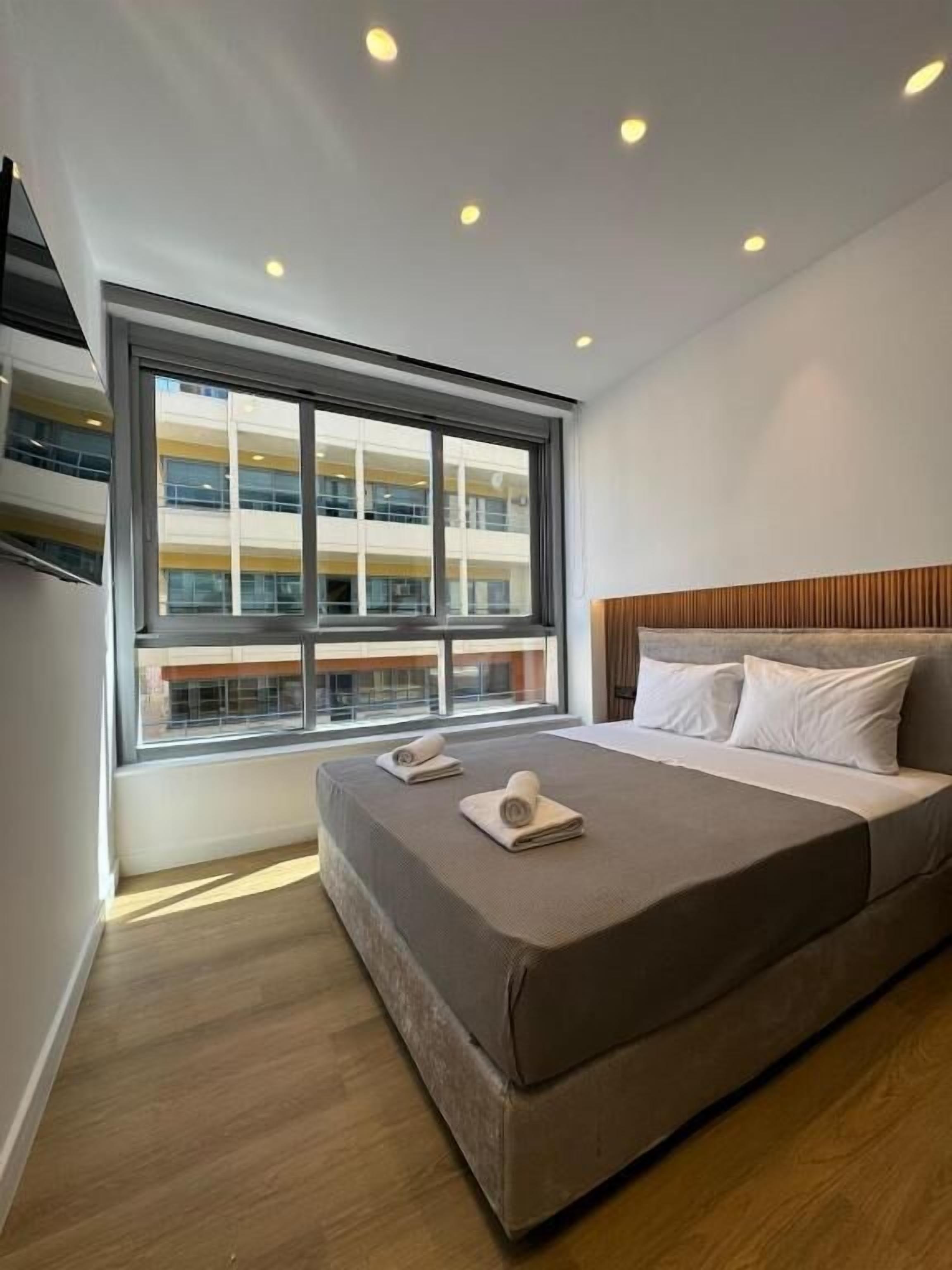A modern hotel room with a large bed, a window with a view of a building, and recessed ceiling lights.