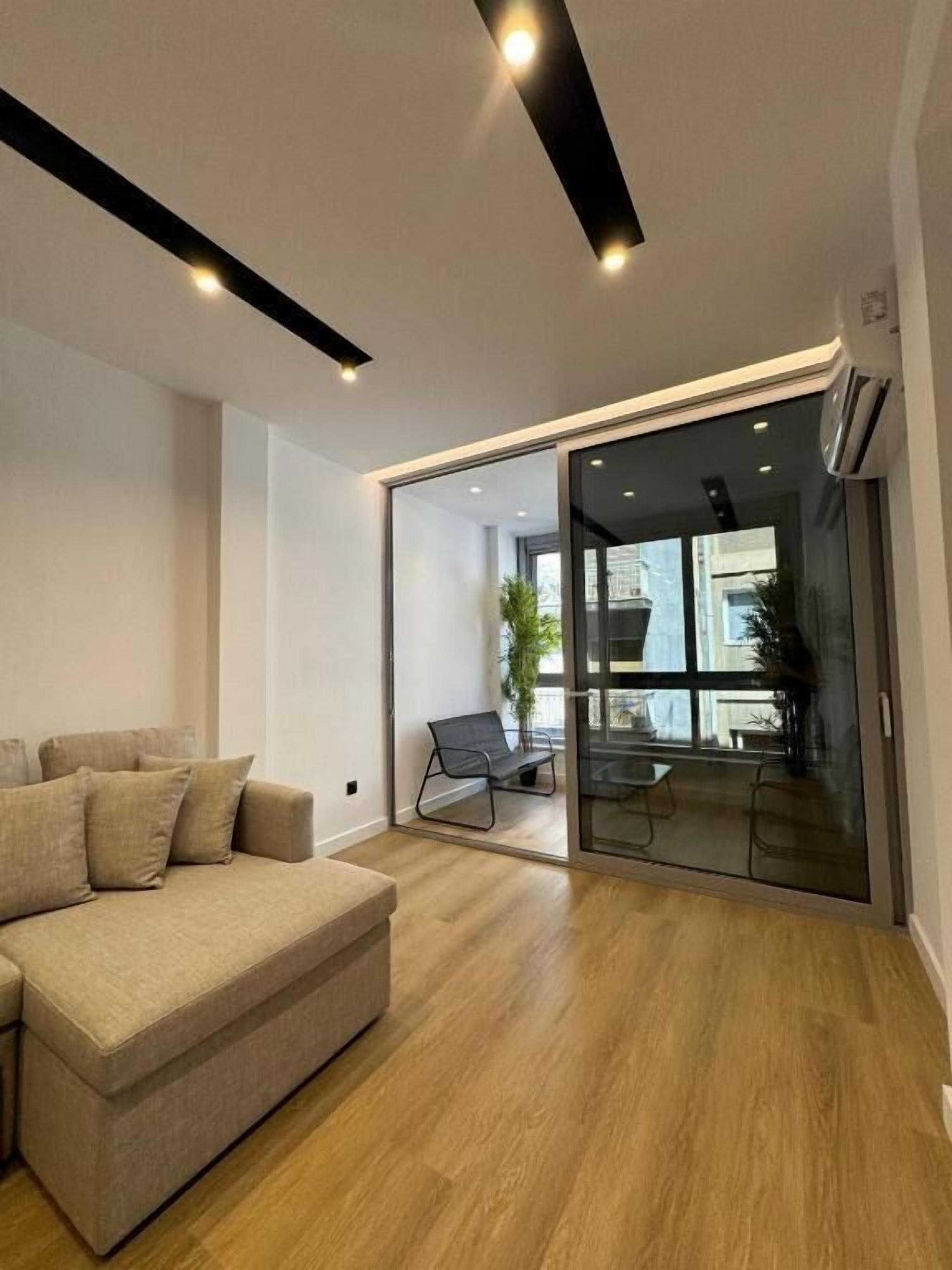 A modern living room with a sofa, a glass door leading to an outdoor area with a chair and table, and a ceiling with recessed lighting.