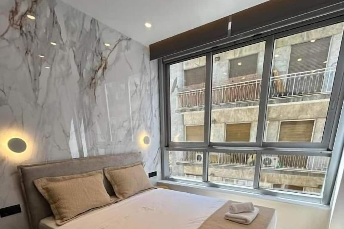 A modern bedroom with a large bed, marble wall, and a view of a building through a large window.