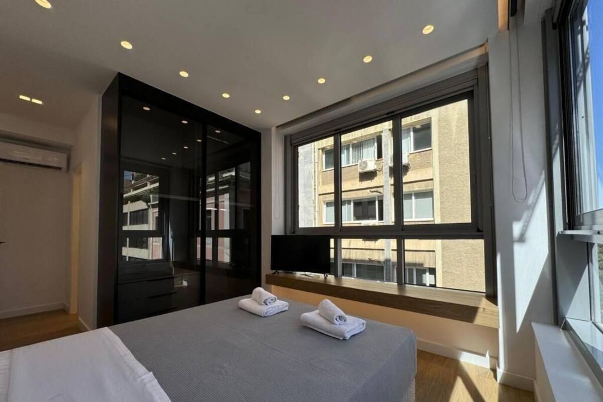 A modern hotel room with a large bed, a television, a window with a view of buildings, and a sliding door closet.