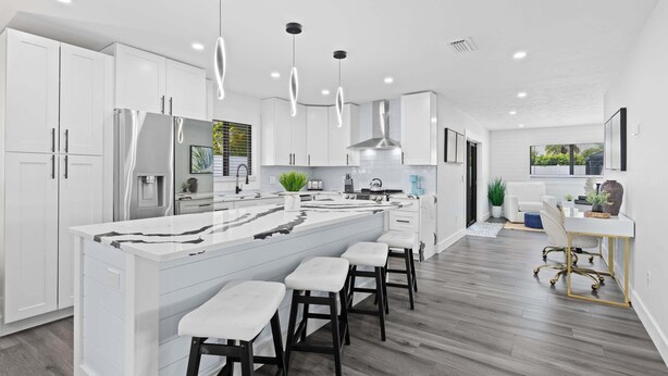 Une cuisine moderne avec des meubles blancs, un îlot central doté de tabourets, un éclairage suspendu et une vue sur l’espace de vie.