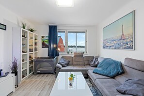 A modern living room with a sofa, a coffee table, a bookshelf, a pet bed, and a large window with a view of a building. - Big one Bedroom Apartment near Airport & City Center (Schwechat)