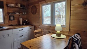 A wooden cabin interior with a kitchen, dining table, chairs, a lamp, and a window with a view of trees.