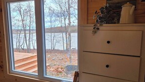 A cabin room with a wooden ceiling, a large window with a view of trees and a dock, a white dresser, and a bed with a quilt.