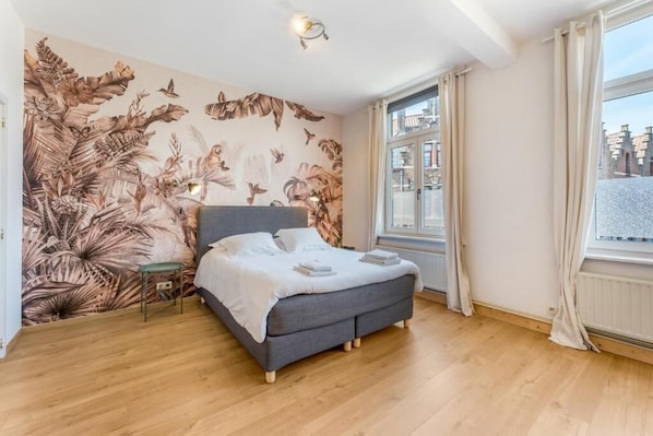 A bedroom with a bed, a wall mural, a window with curtains, and a view of buildings outside. - Werf72 (Bruges)