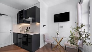 Una cucina moderna con mobili scuri, un piano di lavoro bianco e un forno integrato. È presente un piccolo tavolo rotondo con una pianta e una sedia, e una televisione a schermo piatto fissata alla parete.