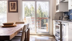 Une cuisine avec une grande fenêtre, une table à manger, des chaises et une vue sur les arbres et une terrasse à l’extérieur.