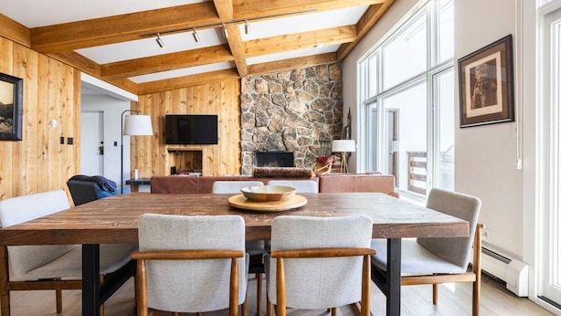 Un vaste espace dédié à la salle à manger, comprenant une grande table en bois, des chaises rembourrées, une cheminée en pierre et un téléviseur.