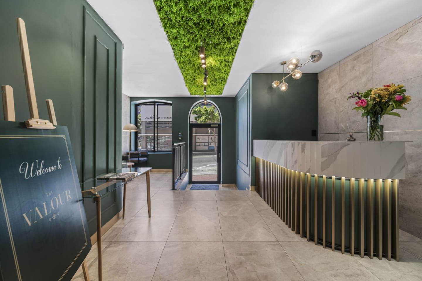 Uma área de recepção com parede verde, balcão de atendimento, espaço para sentar e teto com design que imita grama.