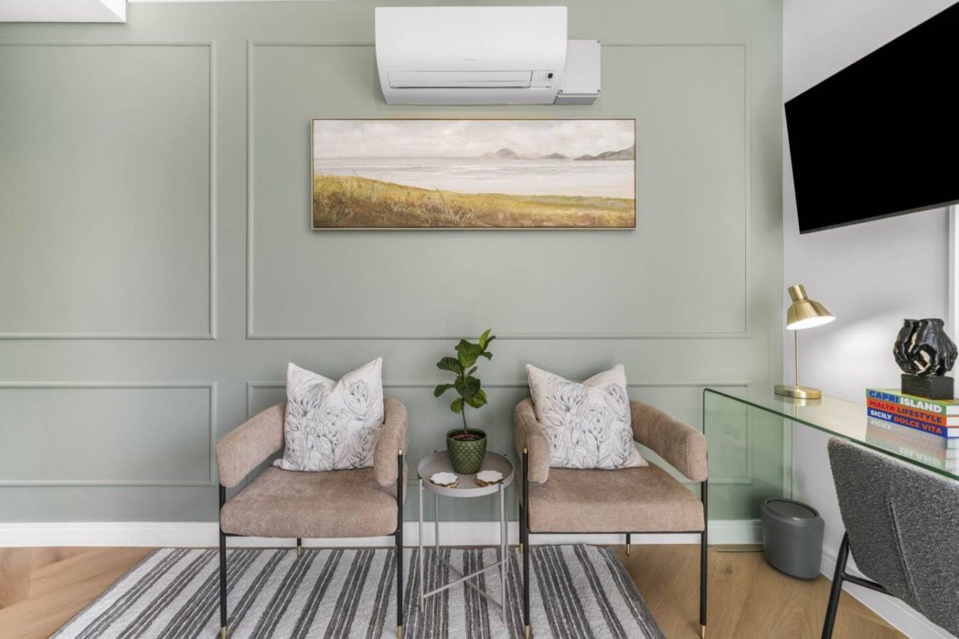 A room with two armchairs, a small table with a plant, a mounted artwork, and a television on the wall.