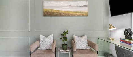 A room with two armchairs, a small table with a plant, a mounted artwork, and a television on the wall.