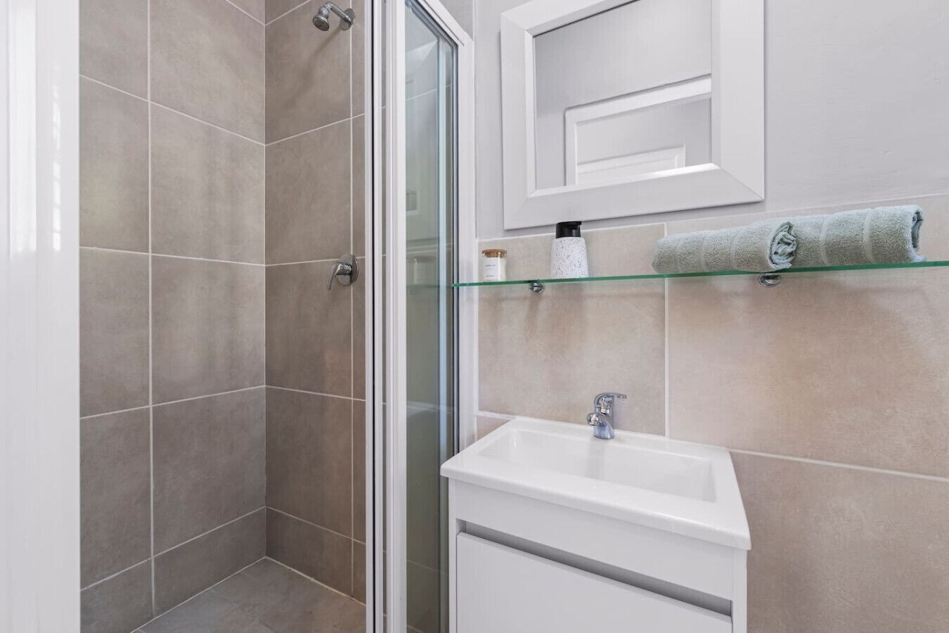 A modern bathroom with a glass shower enclosure, a white sink, and a glass shelf with towels.