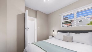 A bedroom with a bed, a nightstand, a lamp, a window, and a door. - Little Daisy Apartment in Tamboerskloof (Cape Town)
