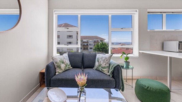A modern living room with a grey sofa, a small table with a vase, and a view of buildings through large windows. - Little Daisy Apartment in Tamboerskloof (Cape Town)