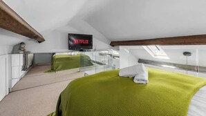 Ein Schlafzimmer mit grüner Bettdecke, einem weißen Kopfkissen und einem Dachfenster. Auf dem Fernseher wird 'NETFLIX' angezeigt.