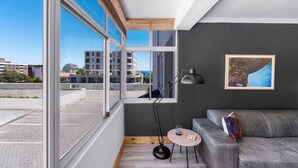 Ein modernes Wohnzimmer mit einem grauen Sofa, einem runden Holztisch, einer Stehlampe und einem großen Fenster mit Blick auf die Stadt.