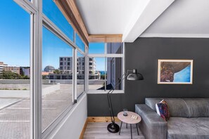 Een moderne woonkamer met een grijze bank, een ronde houten bijzettafel, een vloerlamp en een groot raam met uitzicht op de stads skyline.
