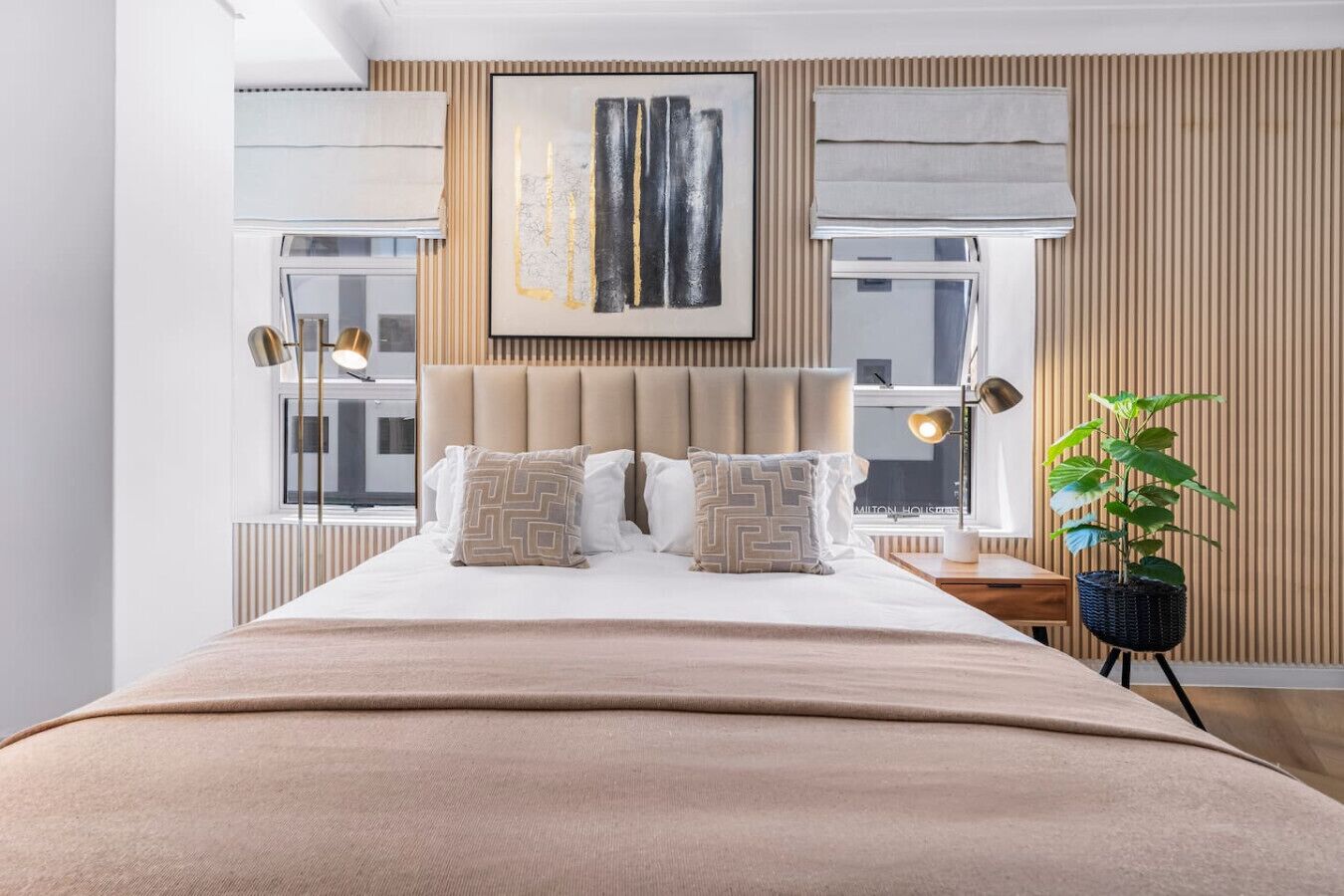 Un dormitorio moderno con una cama grande, un cabecero, una ventana con persianas, una lámpara de noche y una planta.