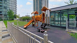 Ein Spielplatz mit Rutsche und Klettergerüsten, umgeben von einem weißen Zaun, in einer städtischen Umgebung mit hohen Gebäuden.