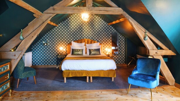 Un dormitorio acogedor en el ático, con techo inclinado, cama con ropa blanca, una silla azul y un aparador de madera.