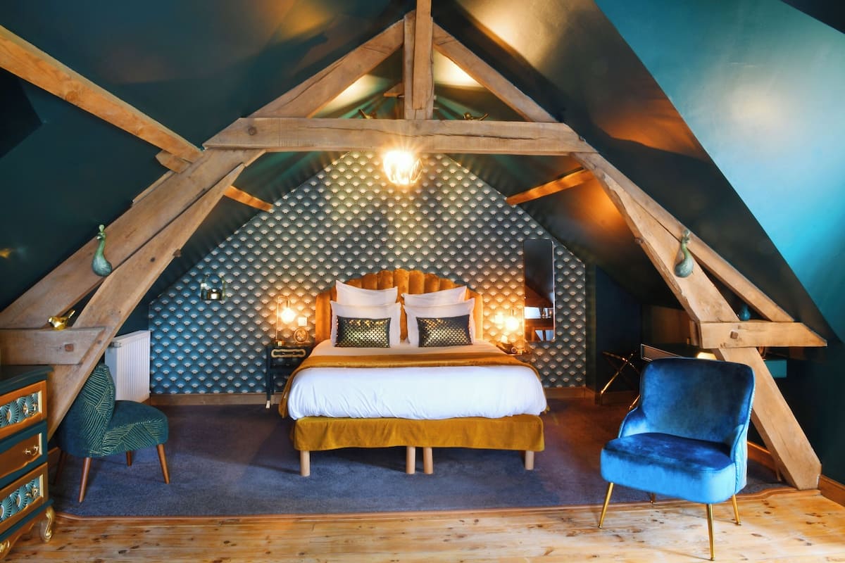 Une chambre mansardée chaleureuse, avec un plafond en pente, un lit recouvert de linge de maison blanc, une chaise bleue et une commode en bois.